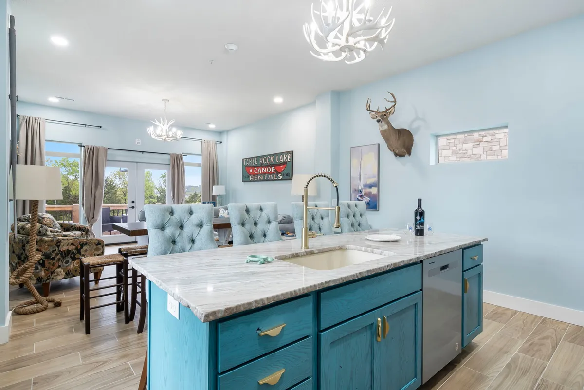 A bright, open kitchen featuring a spacious island with blue cabinets, a stylish chandelier, a mounted deer head, and playful wall signage. The setup encourages both cooking and socializing, with easy flow between prep space and gathering areas.