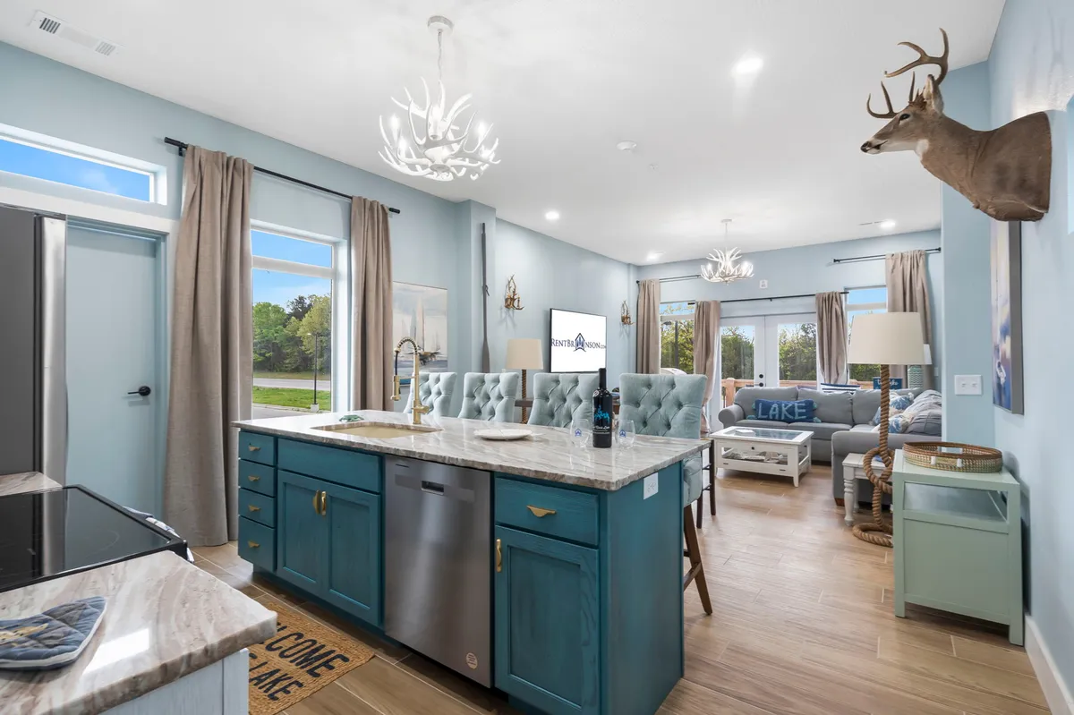 A welcoming, open-concept lounge area featuring a blue kitchen island as the centerpiece, complemented by an antler chandelier, comfortable seating, and decorative accents like a deer mount. The space is designed for both relaxation and entertaining, providing ample room for groups to gather, dine, or play games together.