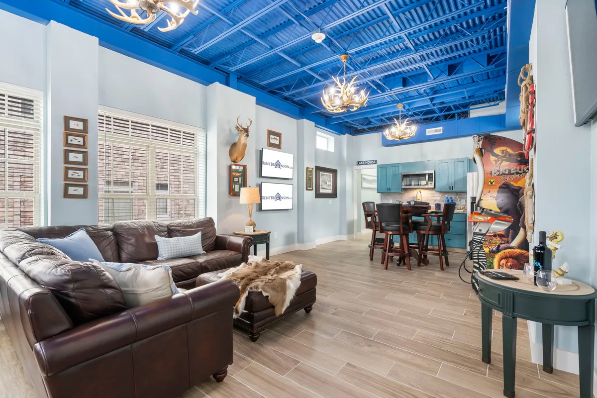 A cozy, lively living area with a leather sofa set against a striking blue ceiling, kitchen space in the background, and decorative wall art. The room is designed for group relaxation and activities, making it perfect for game nights, movie evenings, or casual socializing.