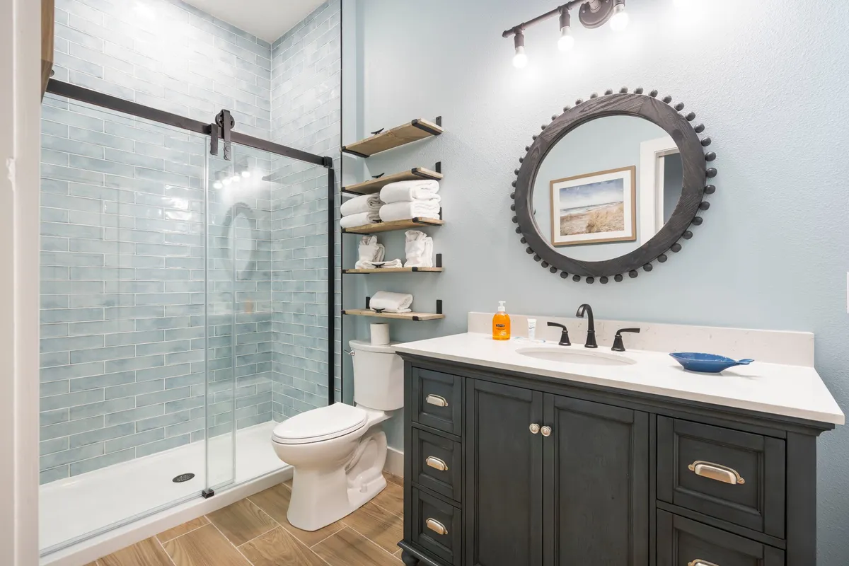 A modern bathroom with a glass shower enclosure, a dark wood vanity, a round decorative mirror, and neatly arranged towels on open shelves. The space feels both inviting and tranquil, ideal for relaxing or refreshing routines.