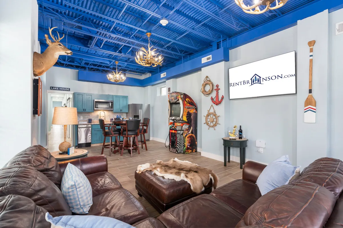 A large leather couch anchors a spacious room with blue ceiling, chandelier lighting, a visible arcade game machine, and cheerful decor including wooden paddles and a mounted deer head. The scene is clearly set up for social gatherings, game nights, and family fun.