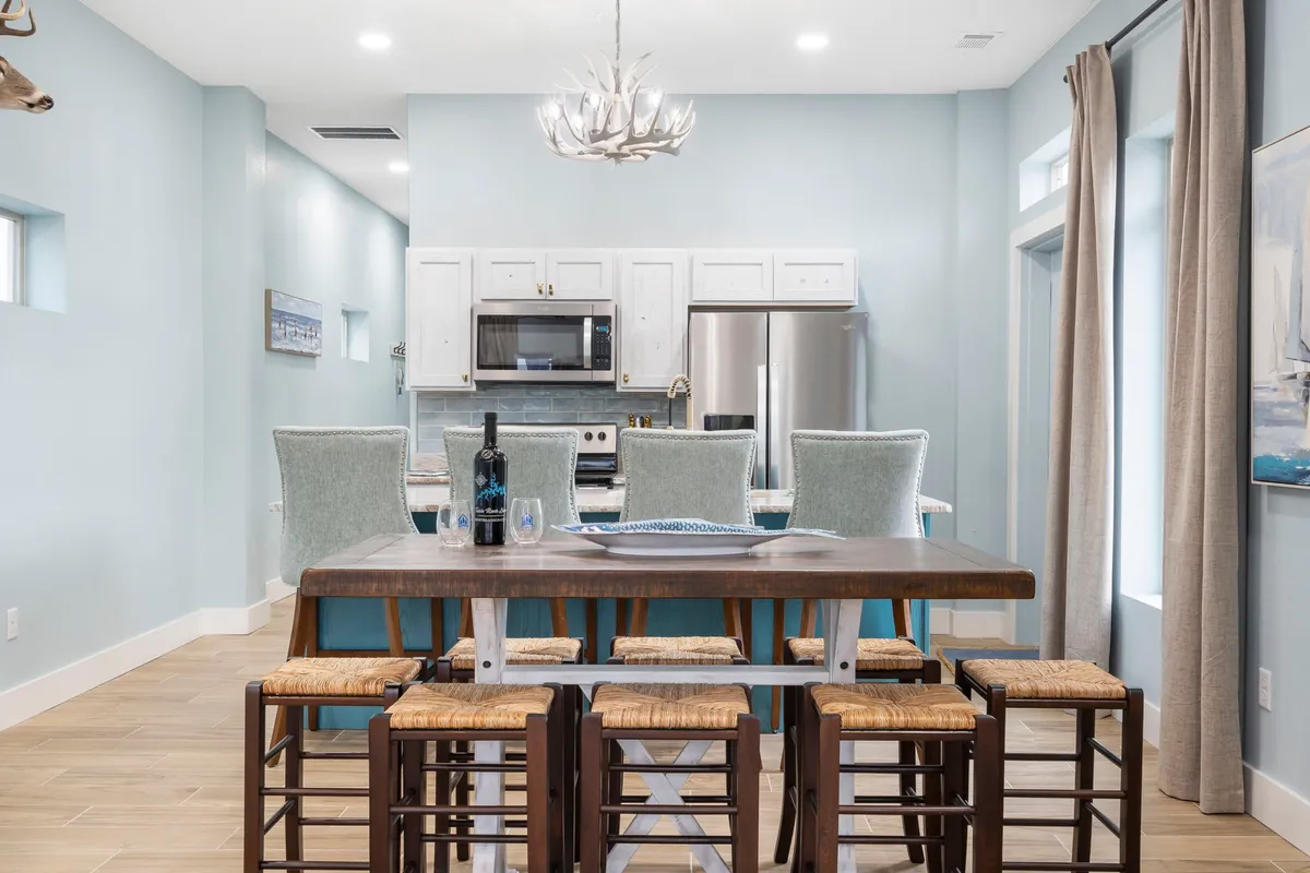 A large wooden dining table surrounded by chairs, adjacent bar stools, and modern kitchen appliances. Warm light from a chandelier creates a cozy, casual setting perfect for family meals and social gatherings.