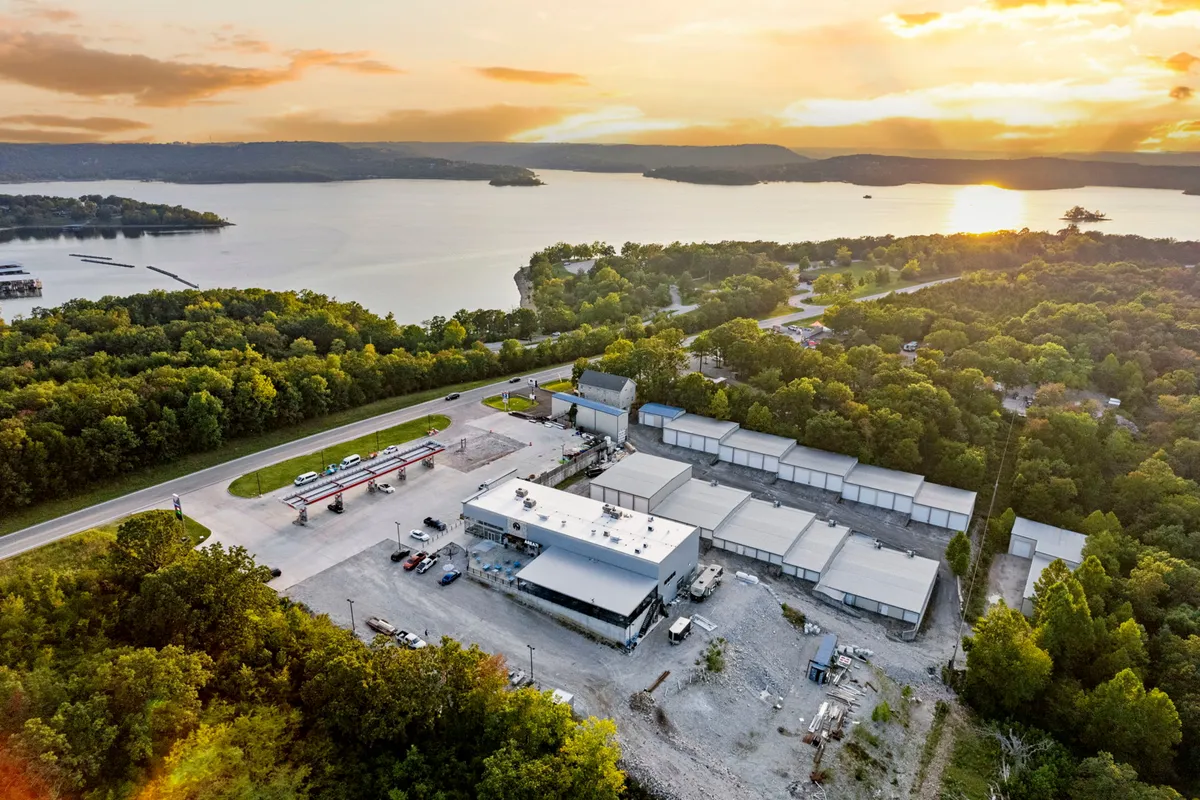 This image presents a serene lake view at sunset, with the tranquil waters of Table Rock Lake reflecting the golden sky. In the background, the marina and an industrial complex are visible, surrounded by lush greenery. People can be seen boating, fishing, and enjoying outdoor activities along the shoreline.