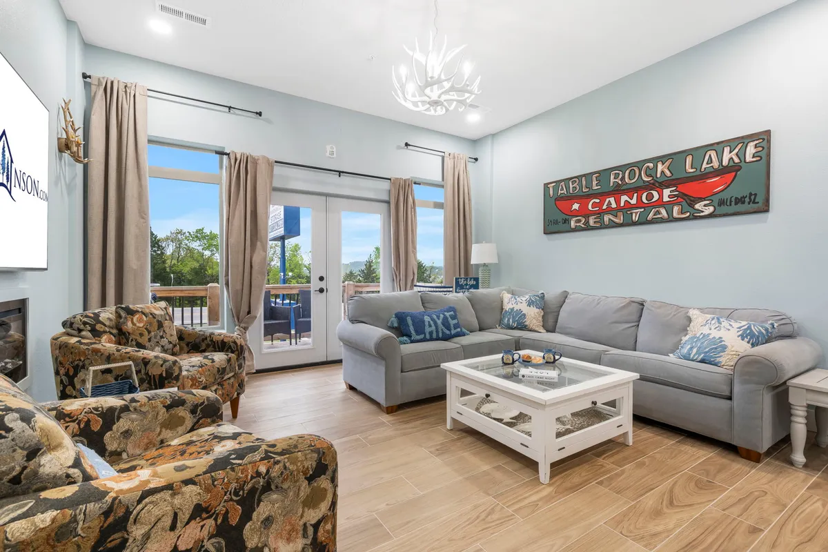 The image showcases a bright, inviting living space with plush seating arranged for conversation, a charming vintage canoe rental sign that nods to lakeside adventures, and expansive windows revealing glimpses of the outdoors. The atmosphere is casual yet stylish, offering a perfect spot for family gatherings, game nights, or enjoying a peaceful sunset view from the deck.