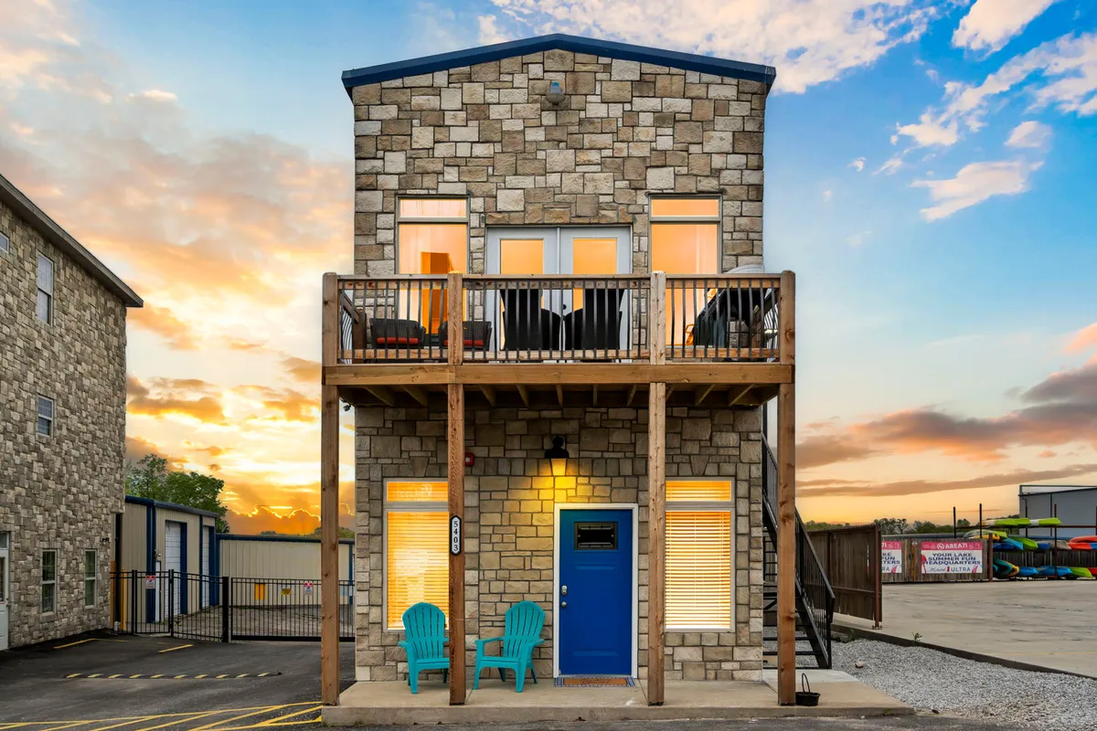 This image shows the exterior stone facade of the property with a blue front door, balcony seating, and a beautiful sunset sky. The inviting balcony hints at the fun and relaxation that awaits inside, and the overall mood is welcoming and lively.