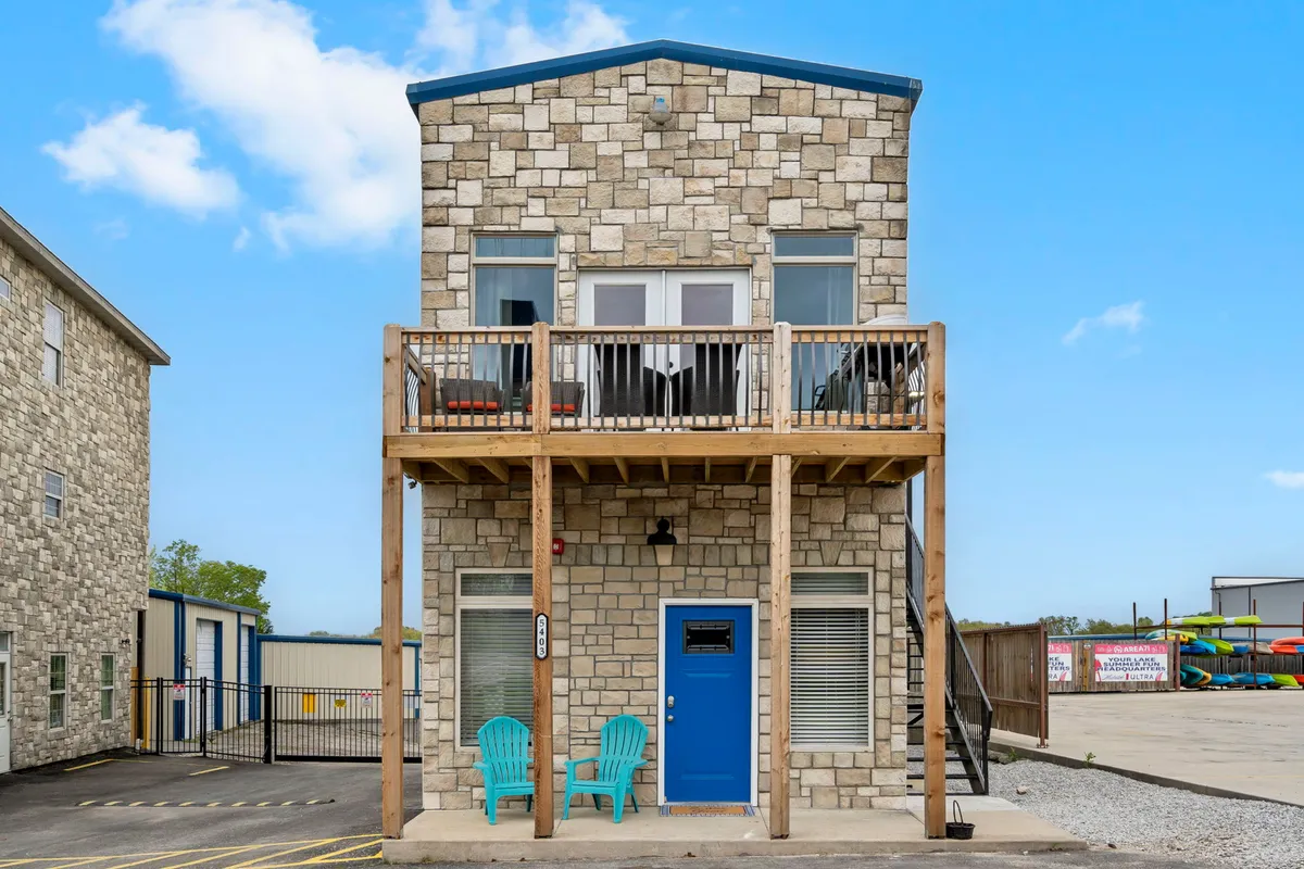 The image features the property's inviting stone facade with a distinctive blue door and a balcony furnished with outdoor chairs. The setting suggests an easy transition from the home to the surrounding outdoor amenities and activities, and hints at enjoying fresh air and neighborhood views.