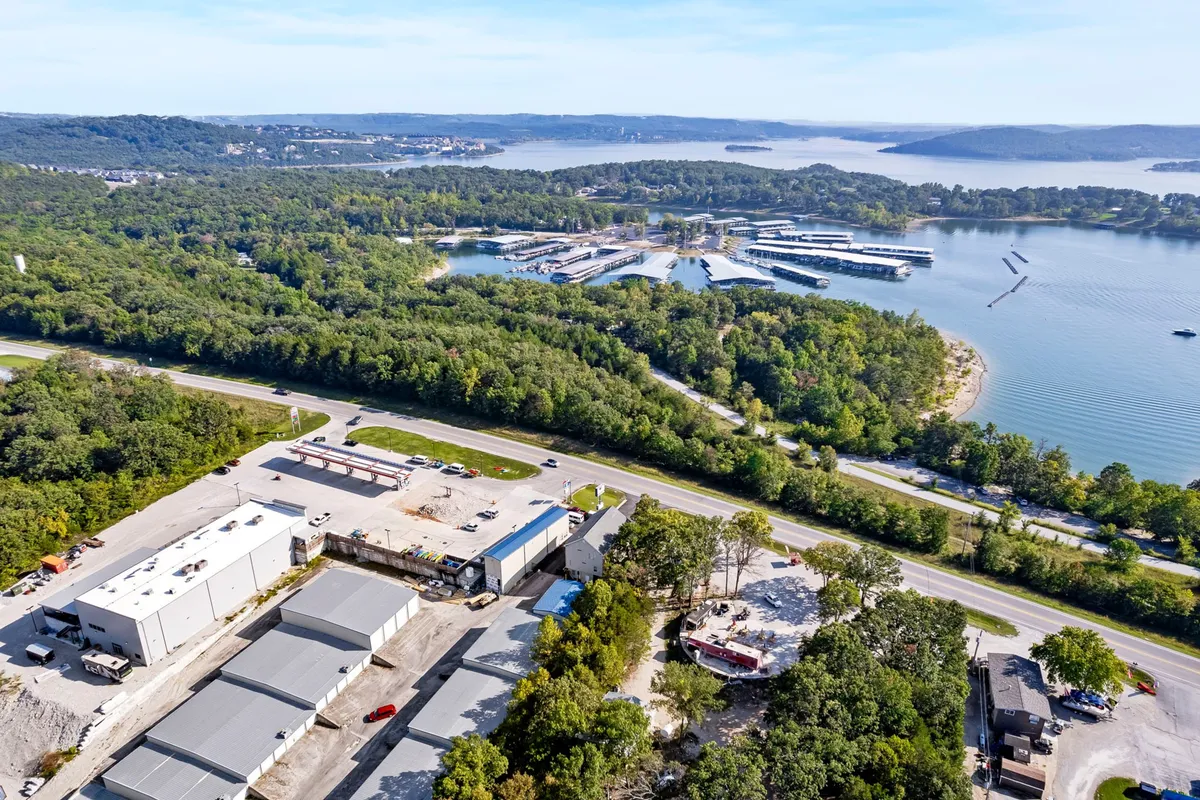 A peaceful marina stretches along the waterfront, framed by a lush forest and calm lake waters. Boats are docked at the marina, and the surrounding area includes a road, some industrial buildings, and inviting spots for fishing or picnicking. The scene is tranquil and inviting, with hints of sunrise or sunset light reflecting off the lake.